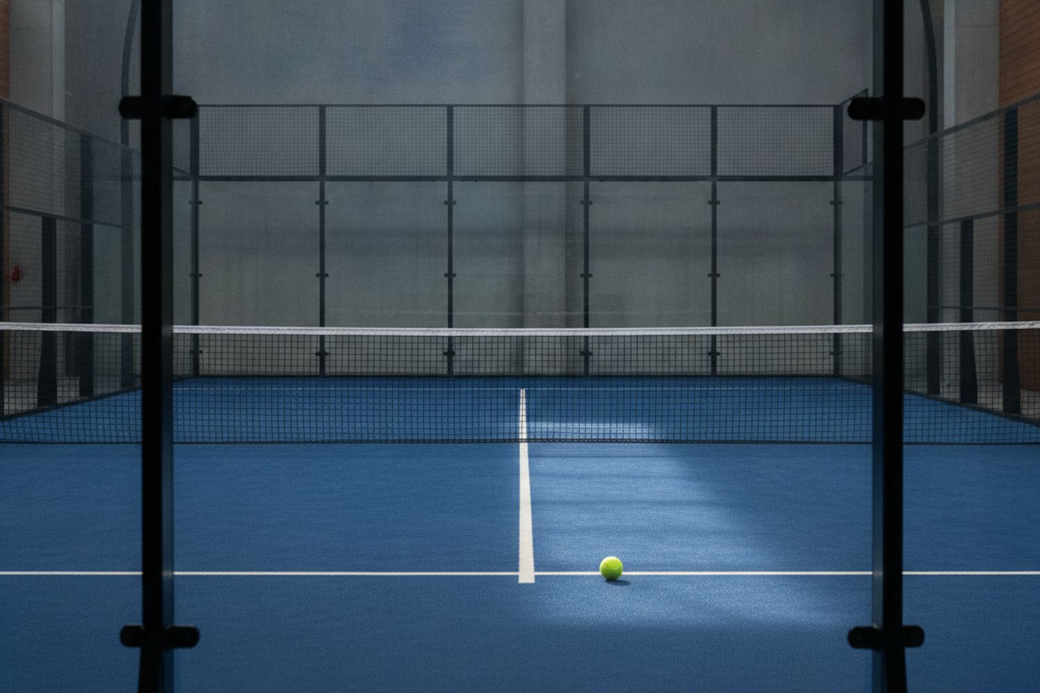The club's blue padel courts behind glass walls at first light, a yellow ball on the centre line